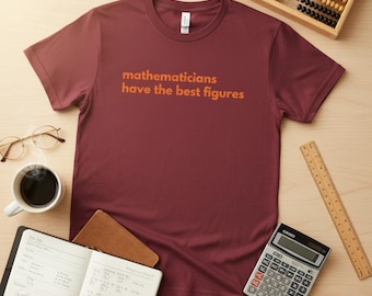 Può includere: Una maglietta bordeaux con la scritta "mathematicians have the best figures" in arancione. La maglietta è circondata da una tazza di caffè, occhiali, un taccuino, una calcolatrice, un righello e un abaco su una superficie di legno chiaro.