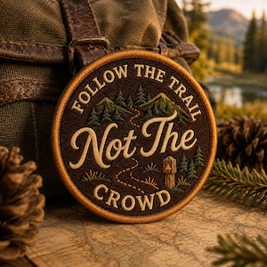 May include: A circular embroidered patch with a dark brown background and a gold border. It features a stitched mountain landscape, a winding trail, and a wooden trail marker. The text reads FOLLOW THE TRAIL Not The CROWD in gold lettering.