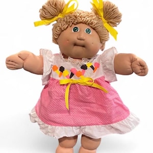 May include: A doll with light brown yarn hair styled in two pigtails with yellow ribbons, green eyes, and dimpled cheeks. The doll wears a white and pink dress featuring a polka dot pattern, a ruffled hem, and a print of colourful ice cream cones.