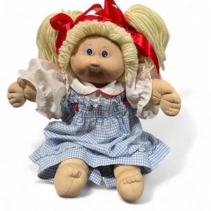 May include: A soft-sculpture doll with blonde yarn hair styled in pigtails tied with red ribbons. The doll has blue eyes, a round face, and wears a white blouse with ruffled sleeves under a blue and white checkered dress with red accents.