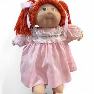 Puede incluir: Una muñeca con cabello de lana naranja peinado en coletas atadas con cintas de cuadros vichy rosa claro y blanco. La muñeca tiene ojos verdes, hoyuelos y viste un vestido rosa claro con cuello de encaje blanco y mangas abullonadas.