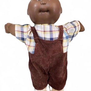 Op de afbeelding: Een pop met een donkerbruine huidskleur en een rond hoofd met bruine ogen en een kleine neus. De pop draagt een shirt met korte mouwen in een wit, blauw en geel ruitpatroon, een donkerbruine ribfluwelen tuinbroek en lichtgekleurde veterschoenen.