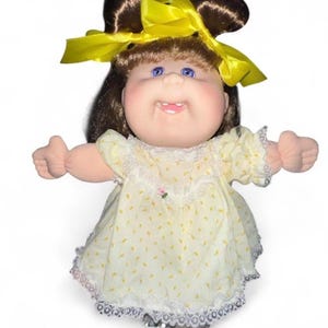 May include: A doll with brown hair styled in two buns, adorned with a bright yellow ribbon. The doll has blue eyes, a small tooth, and wears a pale yellow dress featuring a subtle pattern, white lace trim, and a tiny pink floral accent at the neck.