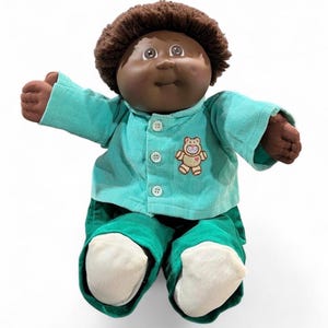 May include: A plush doll with dark brown yarn hair and a soft vinyl face. The doll wears a light teal corduroy long-sleeve shirt with three white buttons and an embroidered teddy bear patch, paired with dark green corduroy pants and white socks.