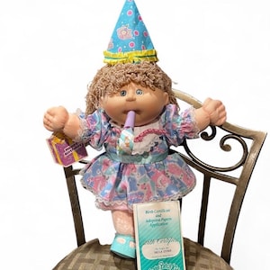 Pode incluir: Uma boneca Cabbage Patch Kids com cabelo castanho encaracolado, usando um chapéu de festa azul com círculos cor-de-rosa e um rebordo amarelo. A boneca veste um vestido azul com um padrão colorido, um colarinho de renda branca e segura um apito de festa roxo. Uma etiqueta diz Happy Birthday e um documento está rotulado como Birth Certificate and Adoption Papers Application.