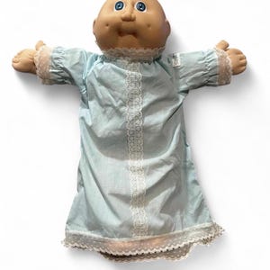 Könnte beinhalten: Eine Puppe mit einem runden, hellbraunen Kopf, Grübchen an den Wangen und leuchtend blauen Augen. Die Puppe trägt ein hellblaues Nachthemd mit einem vertikalen Streifen aus weißer Spitze in der Mitte, Spitzenbesatz an den Ärmeln und einem gerüschten Saum.