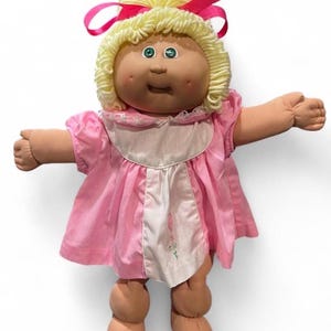 Op de afbeelding: Een Cabbage Patch Kids pop met een lichte huid, groene ogen en kuiltjes in de wangen. De pop heeft blond garen haar in een hoge paardenstaart met een felroze lint. Het draagt een roze jurk met een wit voorpand voorzien van een kleine bloemenborduursel.