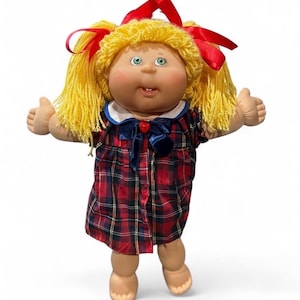 May include: A doll with yarn-like yellow hair styled in pigtails with red ribbons. The doll has green eyes, a small mouth with a visible tooth, and rosy cheeks. It wears a red and navy blue plaid dress with a white collar and a navy bow.