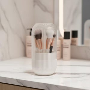 May include: A clear, ribbed plastic makeup brush holder with a white base, containing several makeup brushes with copper-colored handles and soft bristles. The holder sits on a white marble countertop, with blurred bottles in the background.