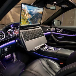 May include: A silver laptop mounted on a steering wheel inside a black car. The laptop screen displays a landscape image. The car interior features black leather seats, ambient purple lighting, and a dashboard with gauges.