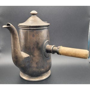 May include: Antique silver-toned coffee pot with a wooden handle. The pot has a curved spout, a lid with a small knob, and a round base. The handle is attached with brass fittings. The pot is likely made of metal and shows signs of age.