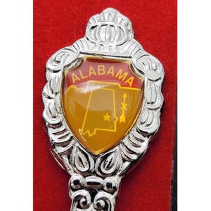 May include: A silver-colored souvenir spoon with a decorative handle. The spoon features a shield-shaped emblem with the word "ALABAMA" and a map of the state in yellow and red. The spoon is set against a red background.