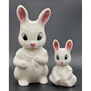 May include: Two white ceramic bunny figurines. The larger bunny has a shawl and the smaller bunny is seated. Both have pink inner ears, red noses, and black eyes. The bunnies are likely decorative items, possibly for Easter.