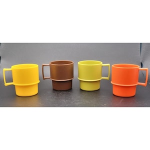 May include: Four colorful plastic mugs in yellow, brown, green, and orange. Each mug has a square handle and a ring around the middle. The mugs are arranged in a row against a dark background.