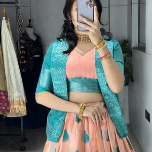 May include: A peach-colored skirt and top set with teal leaf patterns. The outfit includes a teal jacket with a floral design. The person is wearing gold jewelry, including a necklace and bracelets. The outfit is a traditional Indian style.