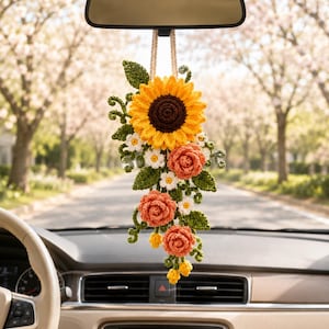May include: A crochet car hanging ornament featuring a large yellow sunflower with a dark brown center, three orange rose-like flowers, small white daisies, and green leaves, all suspended from a beige cord attached to a rearview mirror.