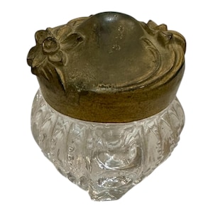 May include: A clear glass jar with a decorative, textured pattern. The jar is topped with a tarnished brass lid featuring floral embellishments. The jar's shape is rounded with a flat bottom.