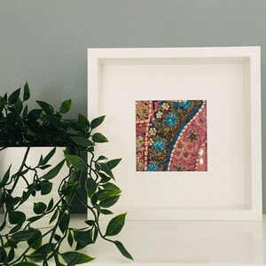 May include: A white frame containing a pink, blue and gold embroidered fabric. The fabric has a floral pattern and is framed with a white mount.