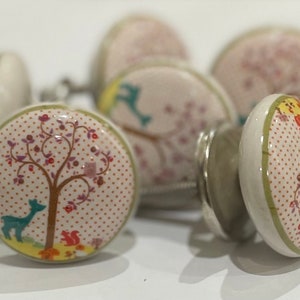 May include: A set of ceramic cabinet knobs with a forest-themed design. Each knob has a cream base with a dotted pattern and a colourful illustration of a tree, deer, and squirrel. The knobs have silver metal hardware.