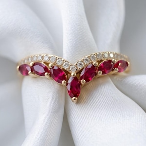 May include: A V-shaped gold ring featuring a row of marquise-cut red rubies and a smaller row of round white stones along the top edge. The band follows a chevron silhouette, showcasing a polished metal finish and vibrant gemstone accents.