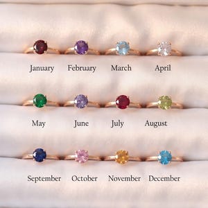 May include: Twelve gold-toned rings with round-cut birthstones arranged in a display case. Each ring is labeled with a month from January to December, featuring stones in red, purple, light blue, clear, green, violet, dark pink, lime, blue, pink, yellow, and cyan.