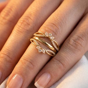 May include: A gold ring set with a unique design. The ring features a curved band with small, sparkling stones and a decorative top with a floral-like arrangement of gemstones. The ring is shown on a finger.