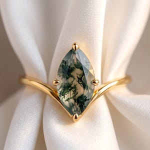 May include: A gold ring featuring a marquise-cut moss agate gemstone with dark green, fern-like inclusions. The stone is held by four prongs on a V-shaped band that tapers toward the center, creating a pointed setting for the faceted, translucent gem.