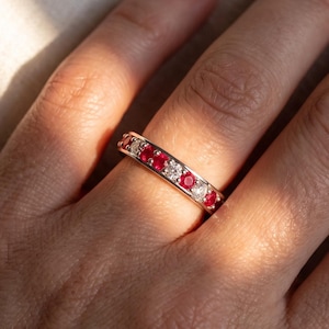 May include: A metal band ring featuring an alternating pattern of round-cut red gemstones and clear white stones set in a continuous row. The ring has a polished finish and is worn on a finger, catching the light to highlight the vibrant red and clear stones.