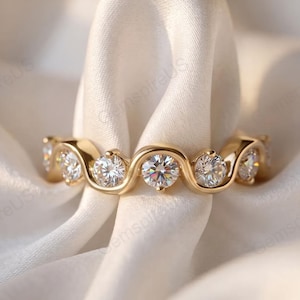 May include: A gold ring featuring a continuous wavy band design. The ring is set with six round, clear, faceted stones arranged in a repeating pattern along the curved metal structure, creating a fluid and interconnected appearance.