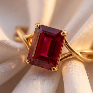 Può includere: Un anello in oro con una grande pietra preziosa rosso scuro dal taglio rettangolare a smeraldo, fissata con quattro griffe. La fascia è realizzata in oro giallo lucido e presenta un design diviso e incrociato che si collega alla pietra.