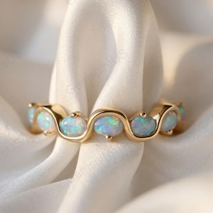 May include: A gold ring featuring a series of oval-shaped opals. The opals have a shimmering, iridescent quality, with flashes of blue, green, and pink. The ring's band has a smooth, polished finish.