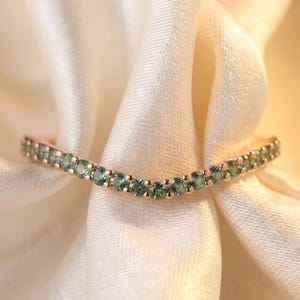 Peut inclure: Une bague en forme de V en métal or rose, ornée d'une série de petites pierres précieuses rondes de couleur verte serties sur toute la longueur de l'anneau. La bague présente une silhouette incurvée en chevron avec une finition métallique.