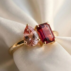 May include: A gold ring with two gemstones: a pear-shaped peach-coloured stone and a rectangular, deep red stone. The ring is set against a backdrop of white fabric, highlighting the ring's design and colours.