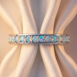 Peut inclure: Une bague en métal argenté ornée d'une rangée continue d'opales rondes et irisées fixées par des griffes. Les pierres présentent des reflets bleus, verts et orange sur leurs surfaces, disposées symétriquement le long de l'anneau métallique.
