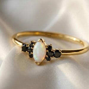 May include: A gold ring with a central oval opal stone, surrounded by clusters of small black stones. The ring band is thin and polished, set against a white, silky background. The ring is a piece of jewelry.