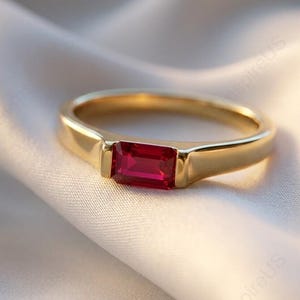May include: A gold ring featuring a rectangular, bright red gemstone. The ring is displayed on a white, silky fabric, accentuating the jewellery's design and colour. The ring's band is a simple, polished gold.