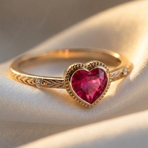 Op de afbeelding: Een gouden ring met een hartvormige, robijnrode edelsteen. De ring heeft een gegraveerde band met kleine diamantaccenten. De hartvormige steen is gezet in een kralenrand, wat zorgt voor een romantisch en elegant ontwerp.