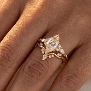May include: A gold engagement ring set featuring a marquise-cut diamond as the centerpiece. The ring is accompanied by a delicate band with smaller accent stones. The design is elegant and classic, perfect for a special occasion.