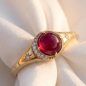 May include: A gold ring featuring a central cabochon-cut red gemstone. The setting includes a crescent-shaped accent adorned with small white stones and engraved star motifs on the band, creating a celestial-inspired design on the metal surface.