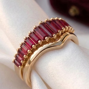 May include: A gold ring set with a row of rectangular, deep red gemstones. The ring is set on a white, silky fabric, with a second ring visible below. A burgundy ring box is in the background.