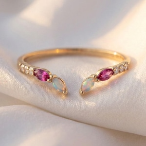 Peut inclure: Une bague dorée avec un design ouvert, ornée de petites pierres claires et de trois pierres précieuses de taille marquise. Les pierres précieuses sont d'un rose vif et d'un blanc nacré. La bague est posée sur un fond blanc texturé.
