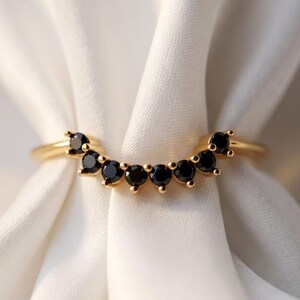 May include: A curved gold ring featuring a row of seven round black stones set in a prong setting. The band has a smooth, polished finish and follows a gentle arc shape, designed to sit flush against another ring. The stones are uniform in size.