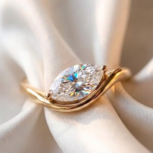 May include: A gold ring featuring a single marquise-cut clear gemstone set horizontally. The band has a smooth, curved, and polished finish that wraps around the base of the stone, creating a sleek and continuous metal silhouette.