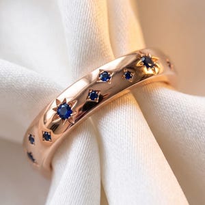 Peut inclure: Une bague anneau en or rose poli ornée de plusieurs petites pierres précieuses rondes bleu foncé incrustées dans le métal. Chaque pierre est entourée d'un motif gravé en forme d'étoile, créant un design céleste sur la surface.