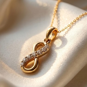 May include: A gold-colored necklace with an infinity symbol pendant. The pendant is adorned with small, clear gemstones. The necklace rests on a cream-colored, silky fabric, creating a luxurious aesthetic. The chain is delicate and complements the pendant's design.