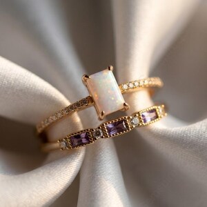 May include: Two gold rings. The top ring features a rectangular, white opal stone. The second ring has a row of small, rectangular purple stones. Both rings have small, clear stones set into the bands.