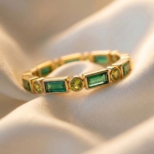 May include: A gold ring featuring alternating emerald-cut and round green gemstones. The ring is set in a gold band and is displayed on a white satin surface. The gemstones are a vibrant green colour.