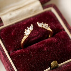 May include: An open-style gold ring featuring two symmetrical ends, each adorned with a cluster of three marquise-cut clear stones arranged in a fan-like pattern. The band is polished gold, showcasing a delicate and refined design.