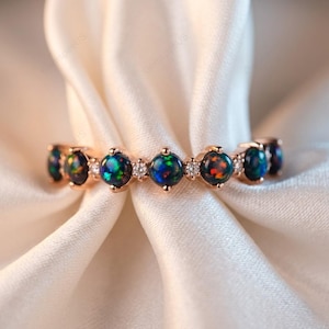 May include: A rose gold ring featuring a series of round, iridescent black opal stones interspersed with small, clear gemstones. The opals display flashes of blue, green, and red. The ring is set against a white, silky fabric.