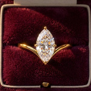 Op de afbeelding: Een gouden ring met een heldere edelsteen in marquise-slijpvorm, gezet in een V-vormige band. De ring is gecentreerd in een dieprode fluwelen doosje, wat de reflecterende facetten van de steen en de gepolijste afwerking van het goud benadrukt.